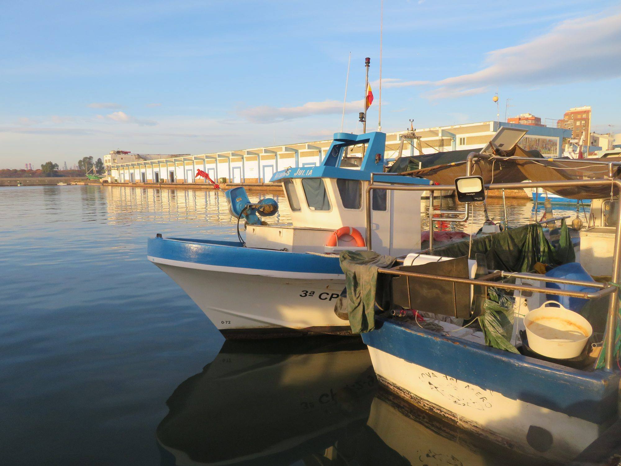 Blue-and-white fishing boats moored in the fishing port of the Grau de Castelló in low, warm sunlight, reflected in calm water, with a long, low white harbour building with blue-framed bays along the quay behind