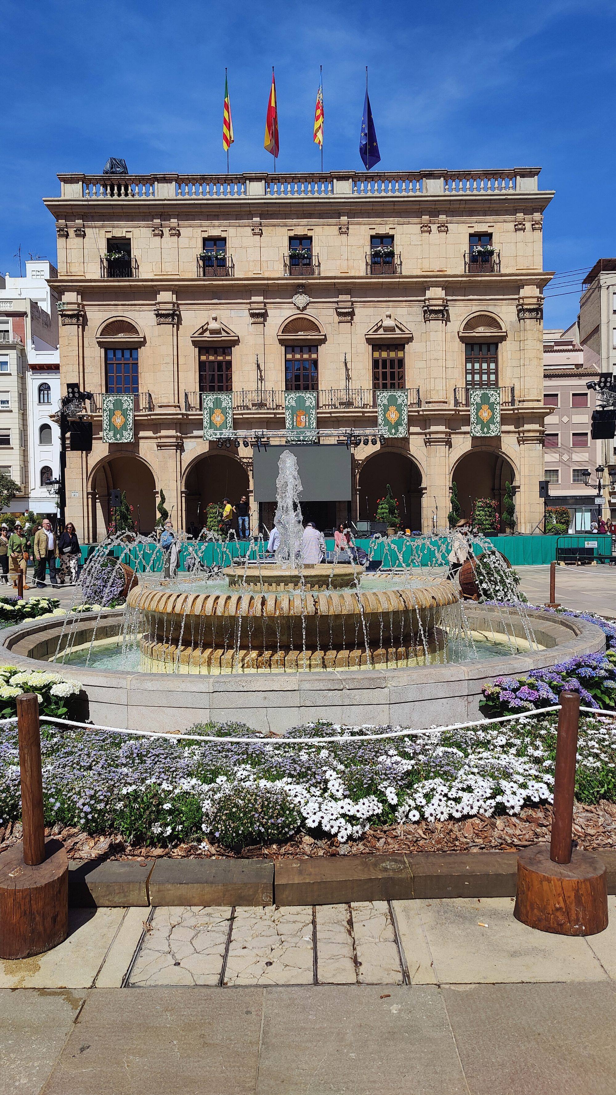 The town hall on the Plaza Mayor of Castellón: a stone façade with an arcaded ground floor, balconies hung with green banners and four flags on the roofline, a temporary stage under the arcades, and in front a round fountain ringed with beds of white and lilac flowers, under a deep blue sky