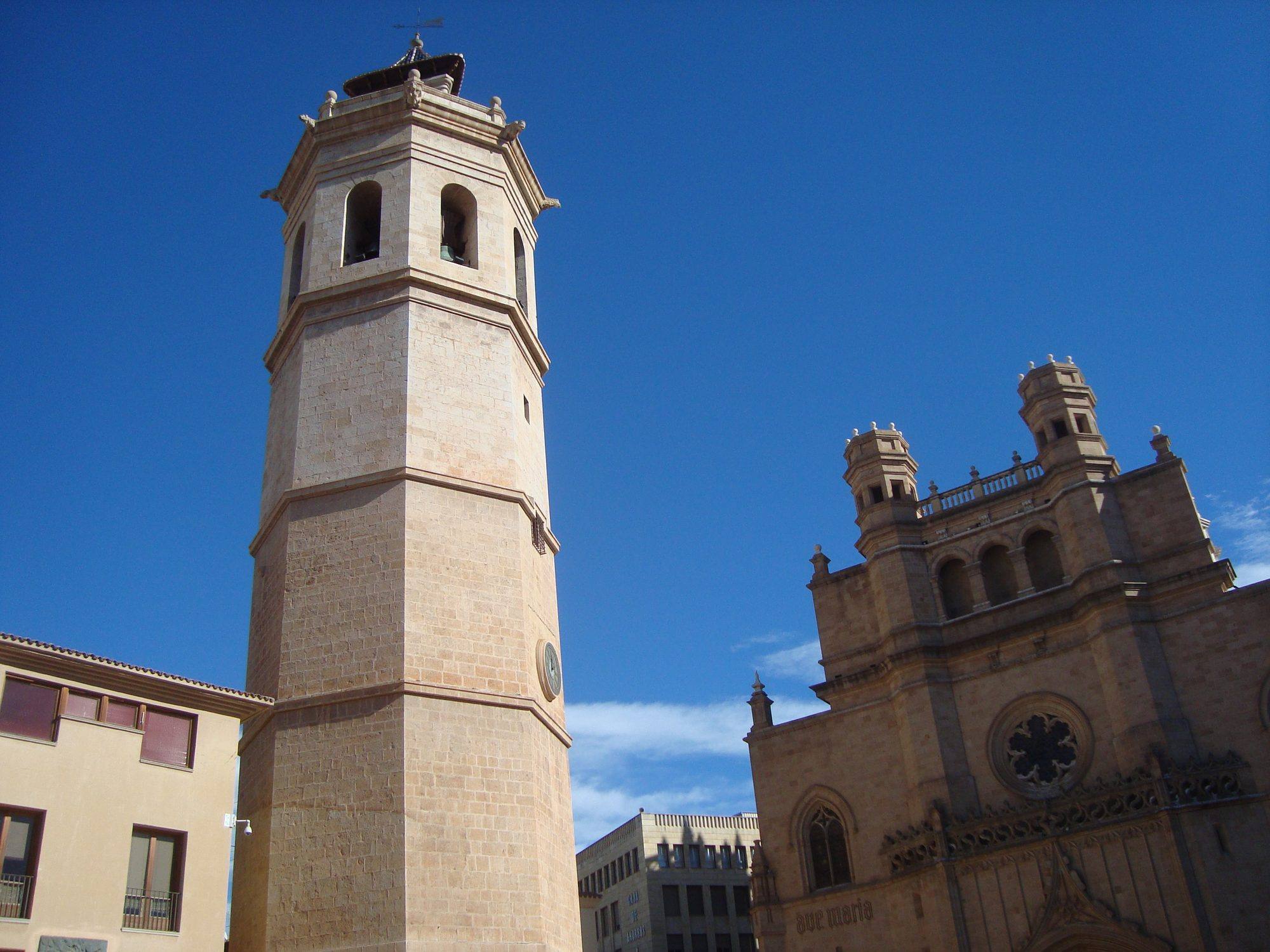 El Fadrí, Castellón's free-standing octagonal stone bell tower, rising into a deep blue sky with its arched bell openings, gargoyles and small tiled roof topped by a weathervane, beside the upper façade of the Concatedral de Santa María on the Plaza Mayor, with its rose window, Gothic tracery frieze and twin octagonal turrets