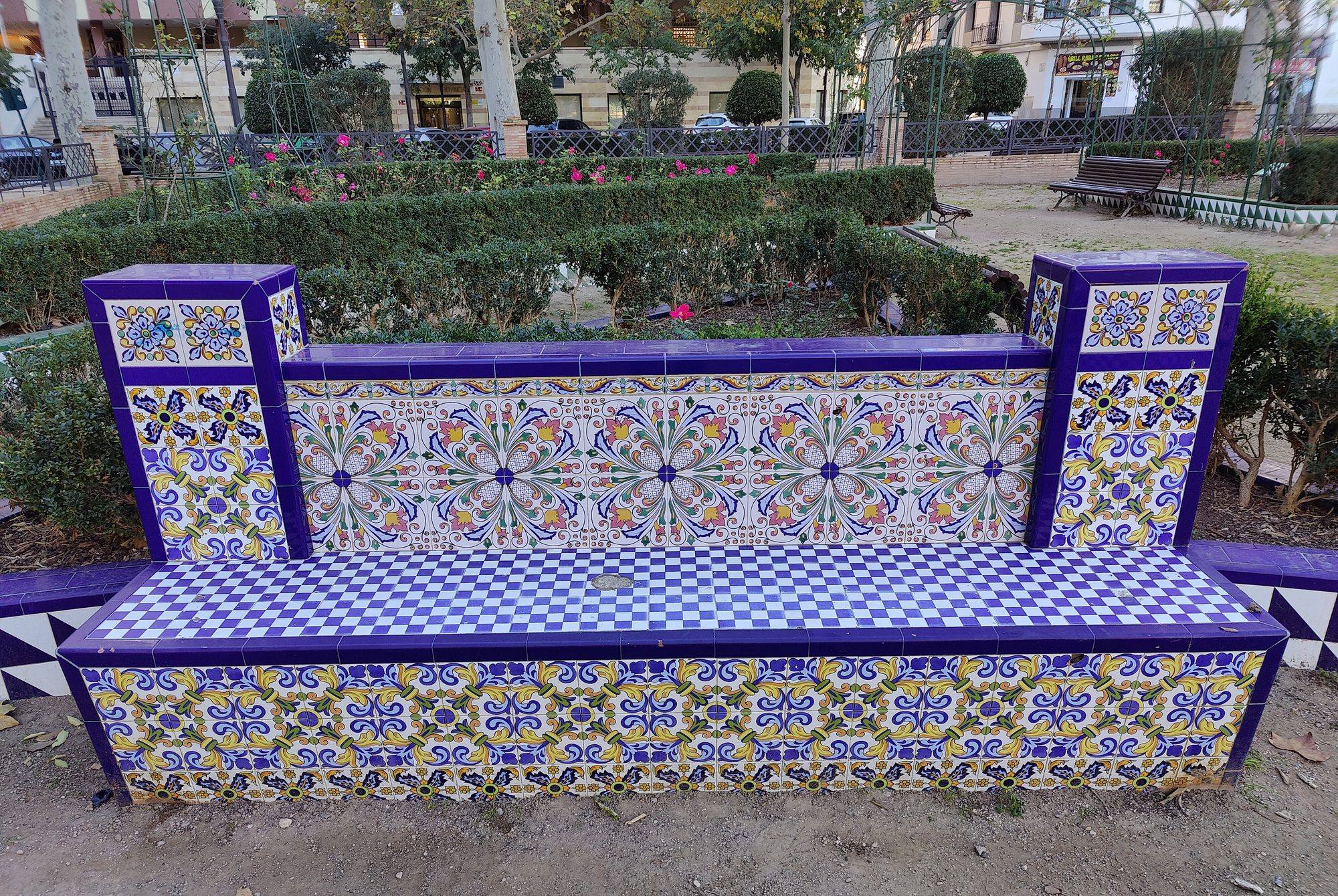 A tiled bench in the rose garden of the Parc Ribalta: the backrest covered in large floral tiles in blue, pink, yellow and green, the seat in a blue-and-white checkerboard, the base and end posts in yellow-and-blue scroll patterns edged with deep blue tiles, in front of clipped hedges and rose bushes
