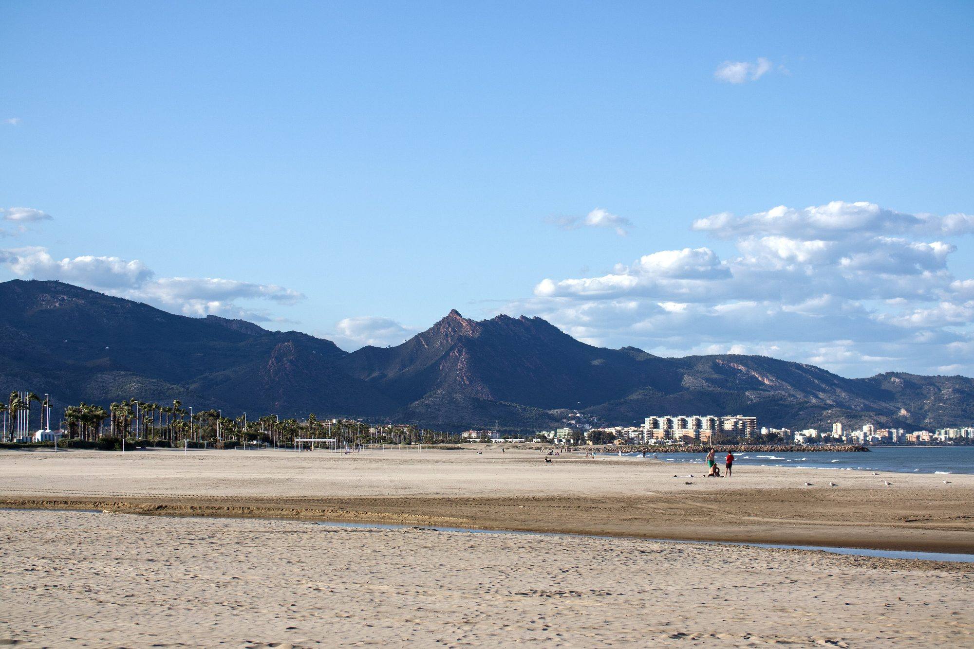 The wide, nearly empty sand of Gurugú beach in Castellón in late-afternoon light, with palms along the promenade, apartment blocks along the shore to the north and the jagged peaks of the Desert de les Palmes behind, under a blue sky with clouds